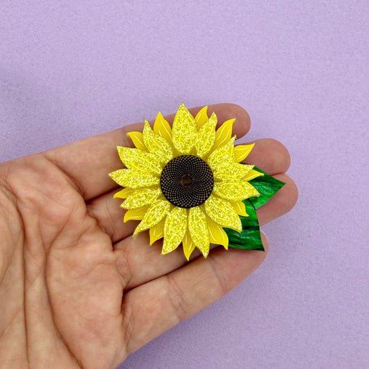 Hand holding a yellow sunflower brooch against a purple background