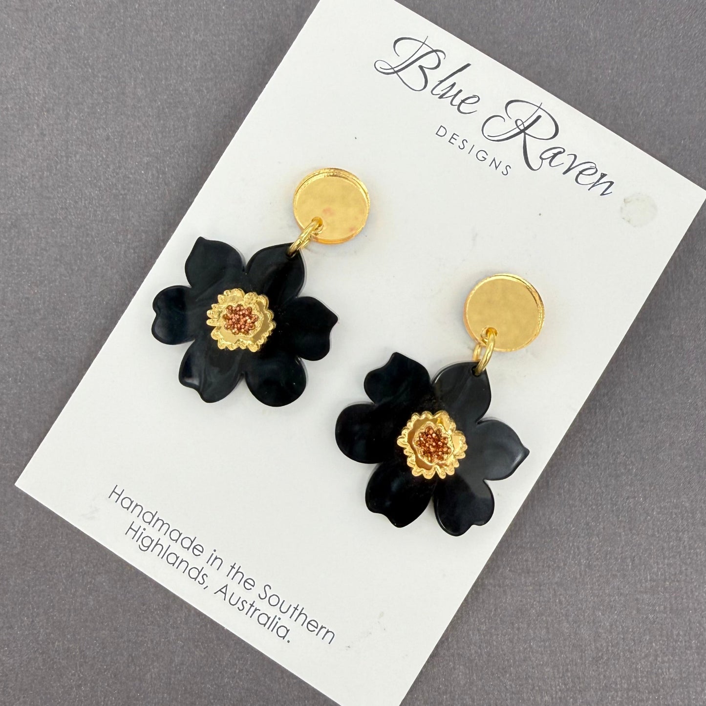 Black flower earrings with gold centers on a Blue Raven Designs card against a gray background