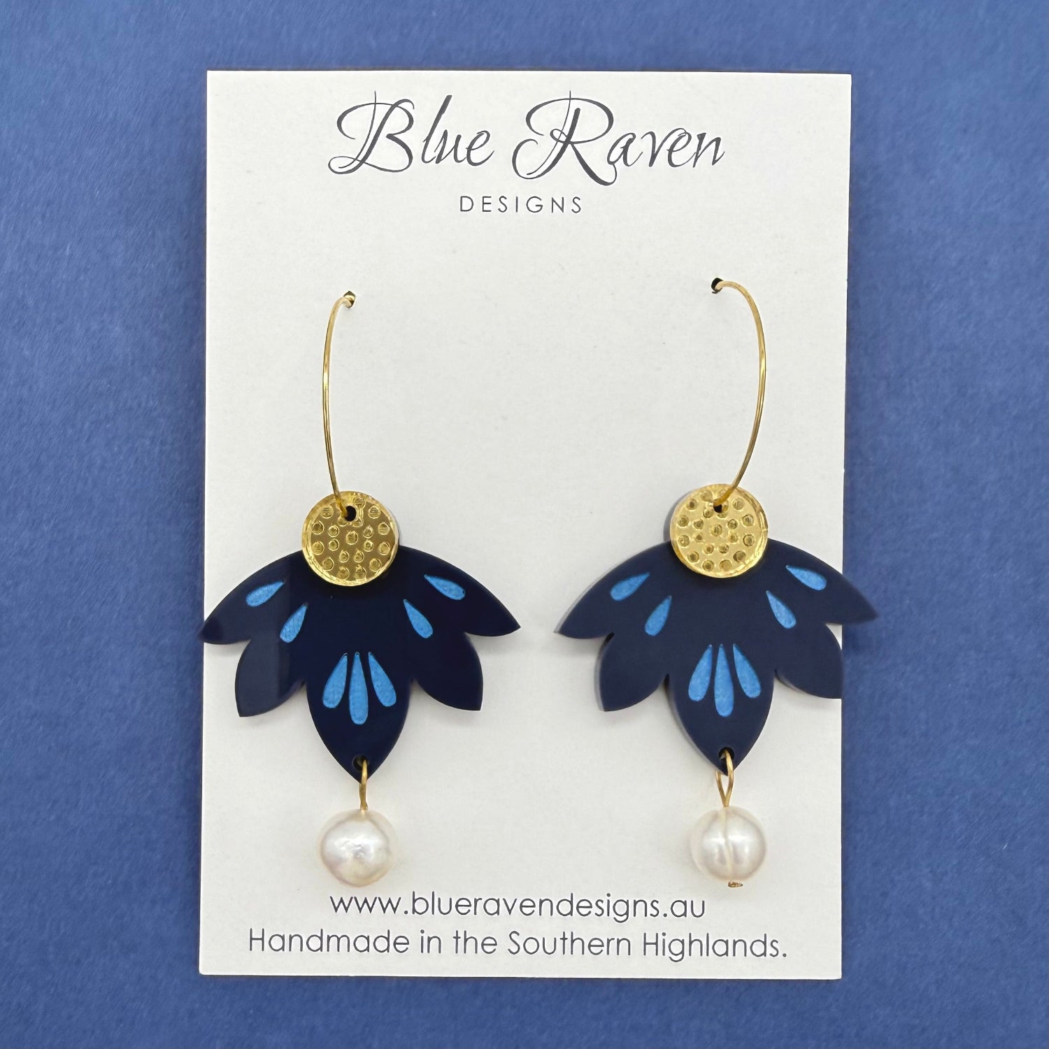 Pair of blue flower-shaped earrings with gold centers and pearl drops on a Blue Raven Designs card against a blue background.