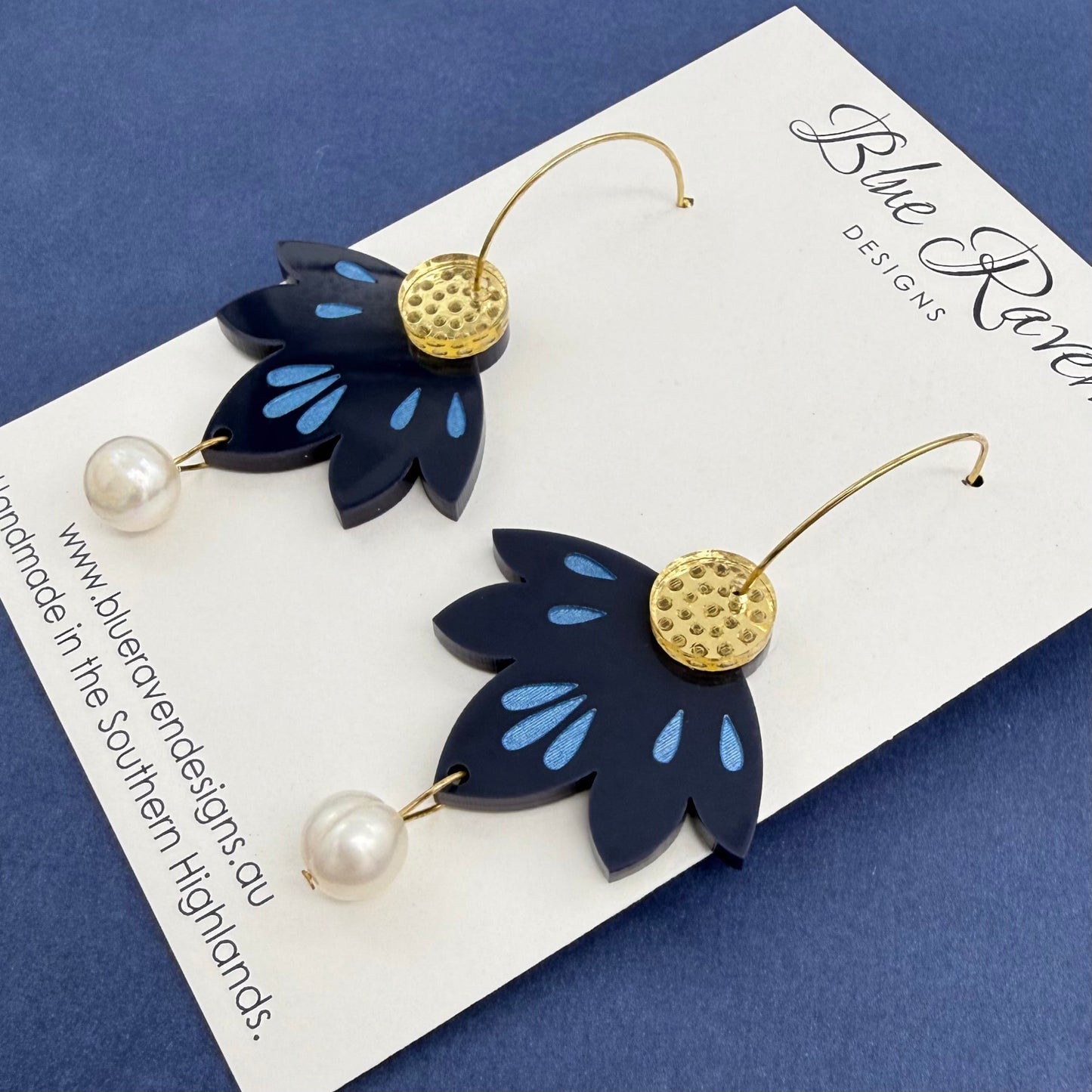 Navy blue leaf-shaped earrings with gold centers and pearl details on a Blue Raven Designs card.