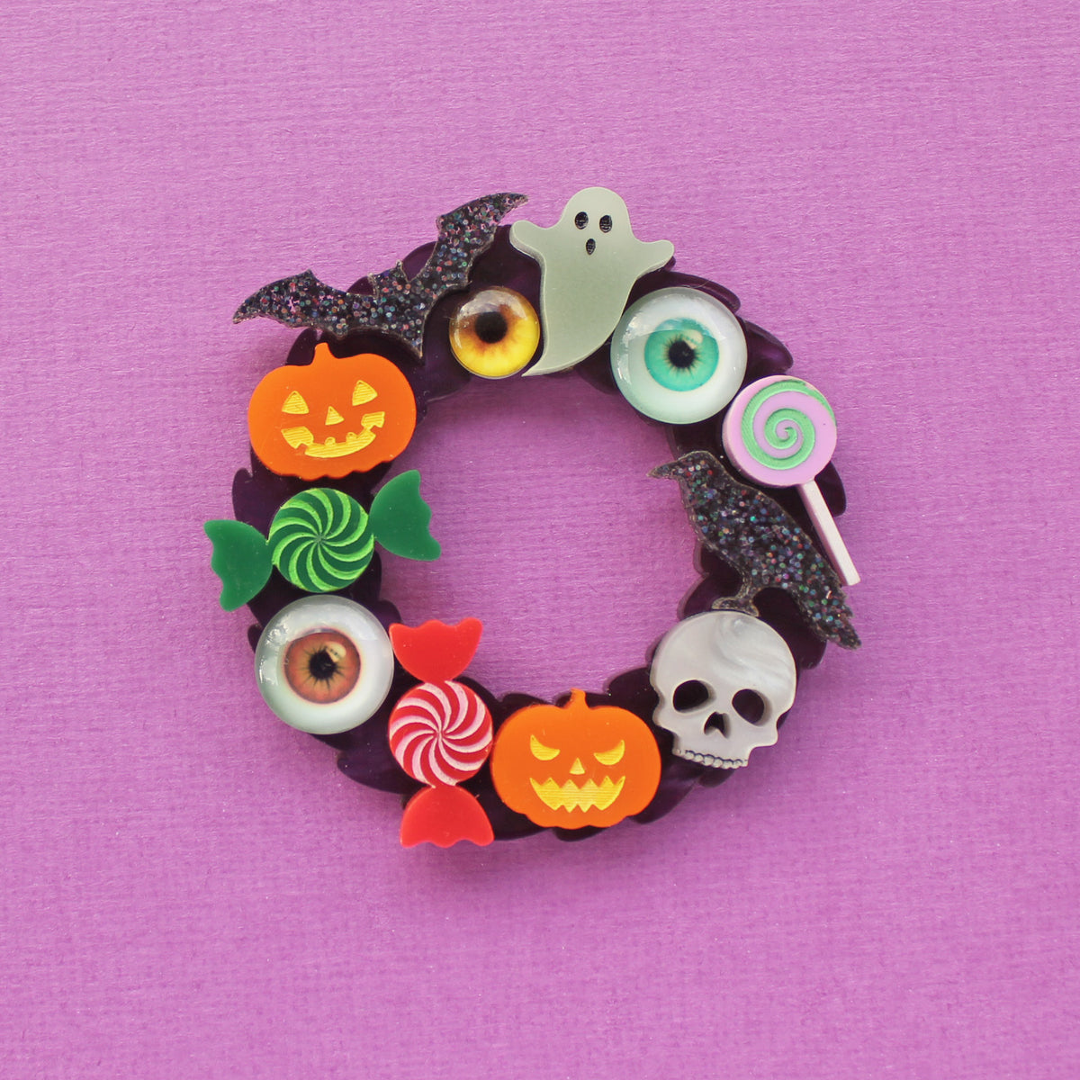 A circular Halloween-themed brooch featuring a dark purple base decorated with candy, lollipops, eyeballs, pumpkins, a ghost, a skull, and bat shapes, displayed on a purple background.