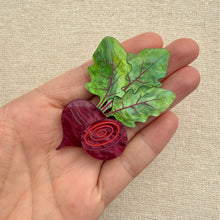 Load image into Gallery viewer, Hand holding an acrylic brooch of a beet with leaves against a beige fabric background.
