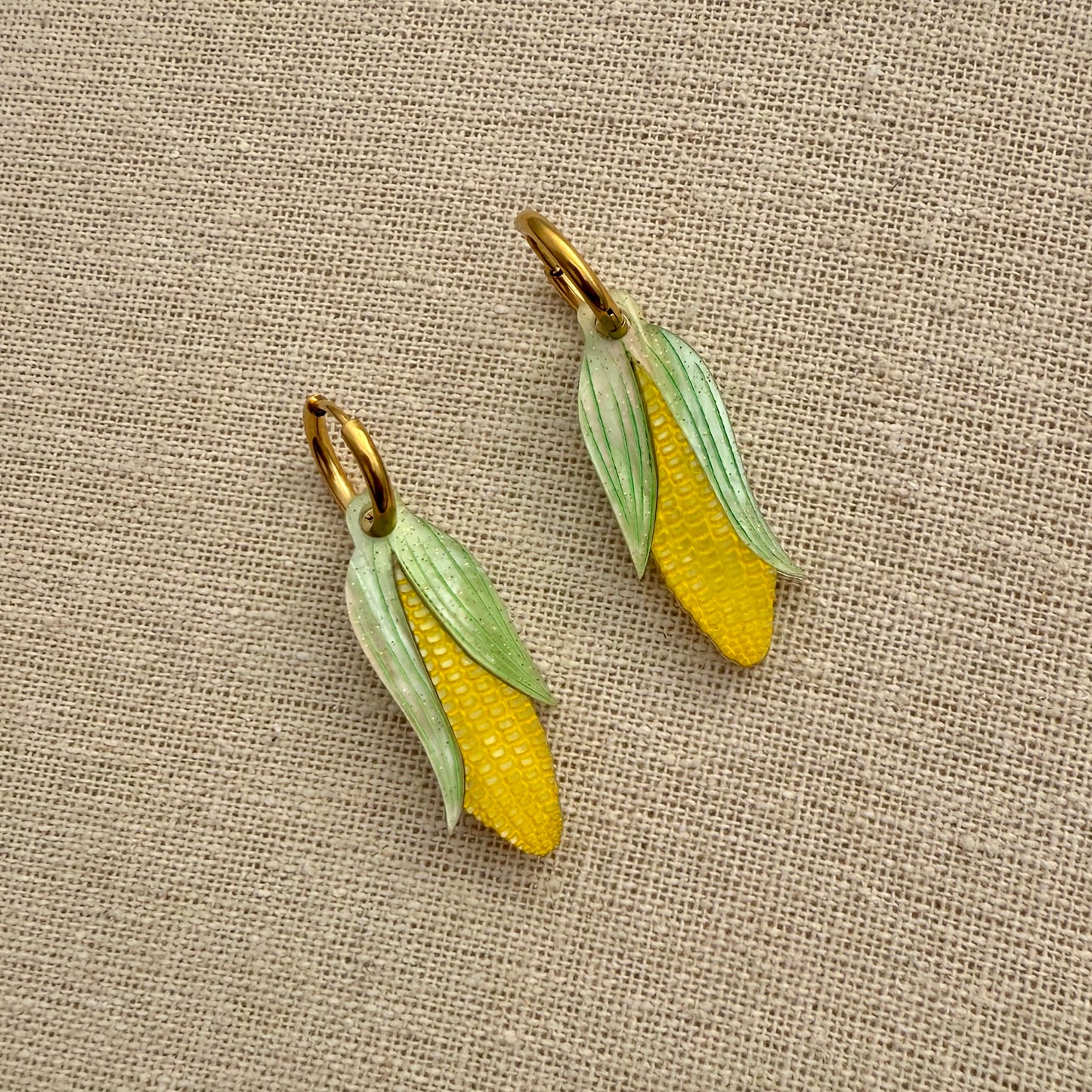 Two corn-shaped earrings on a textured beige fabric background