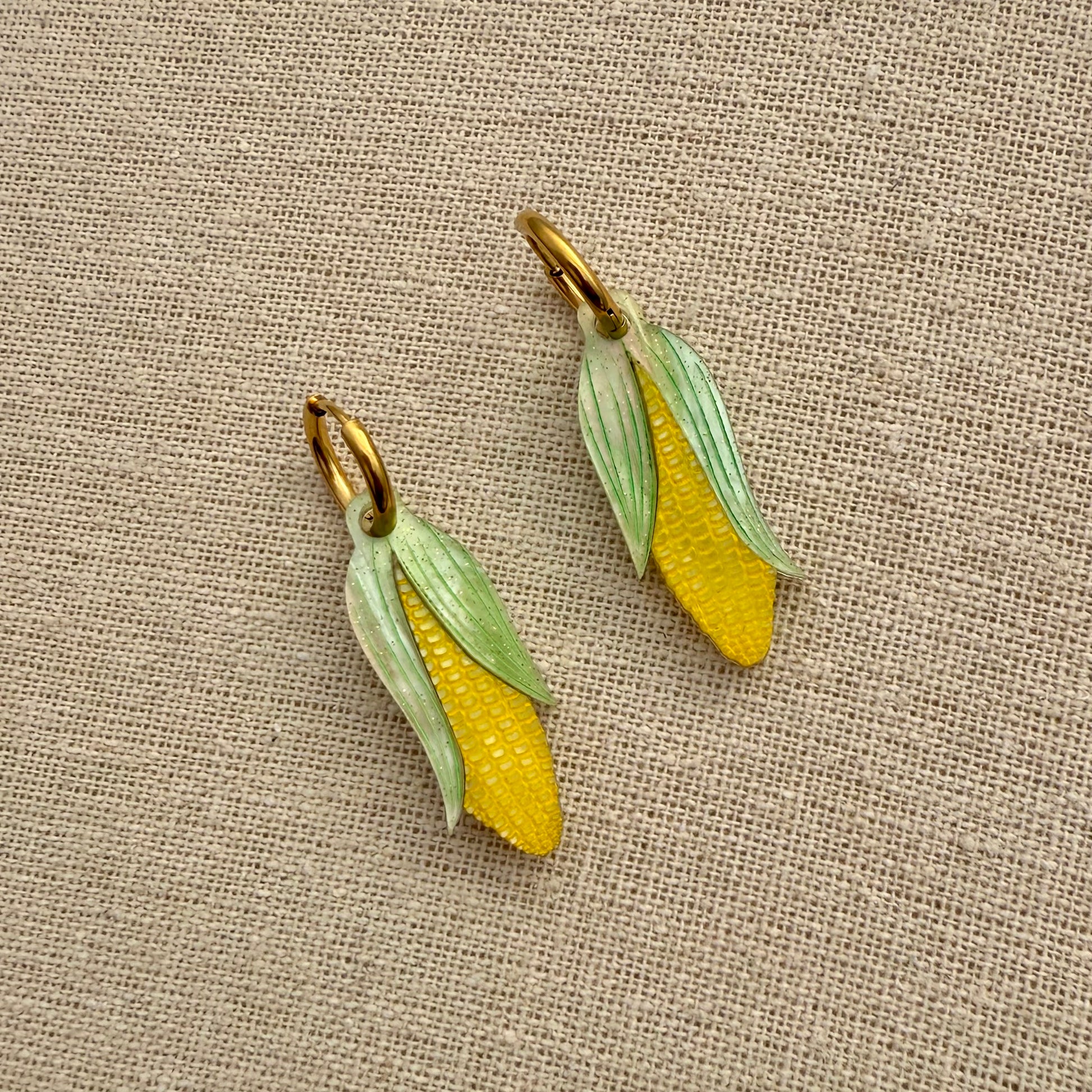 Two corn-shaped earrings on a textured beige fabric background