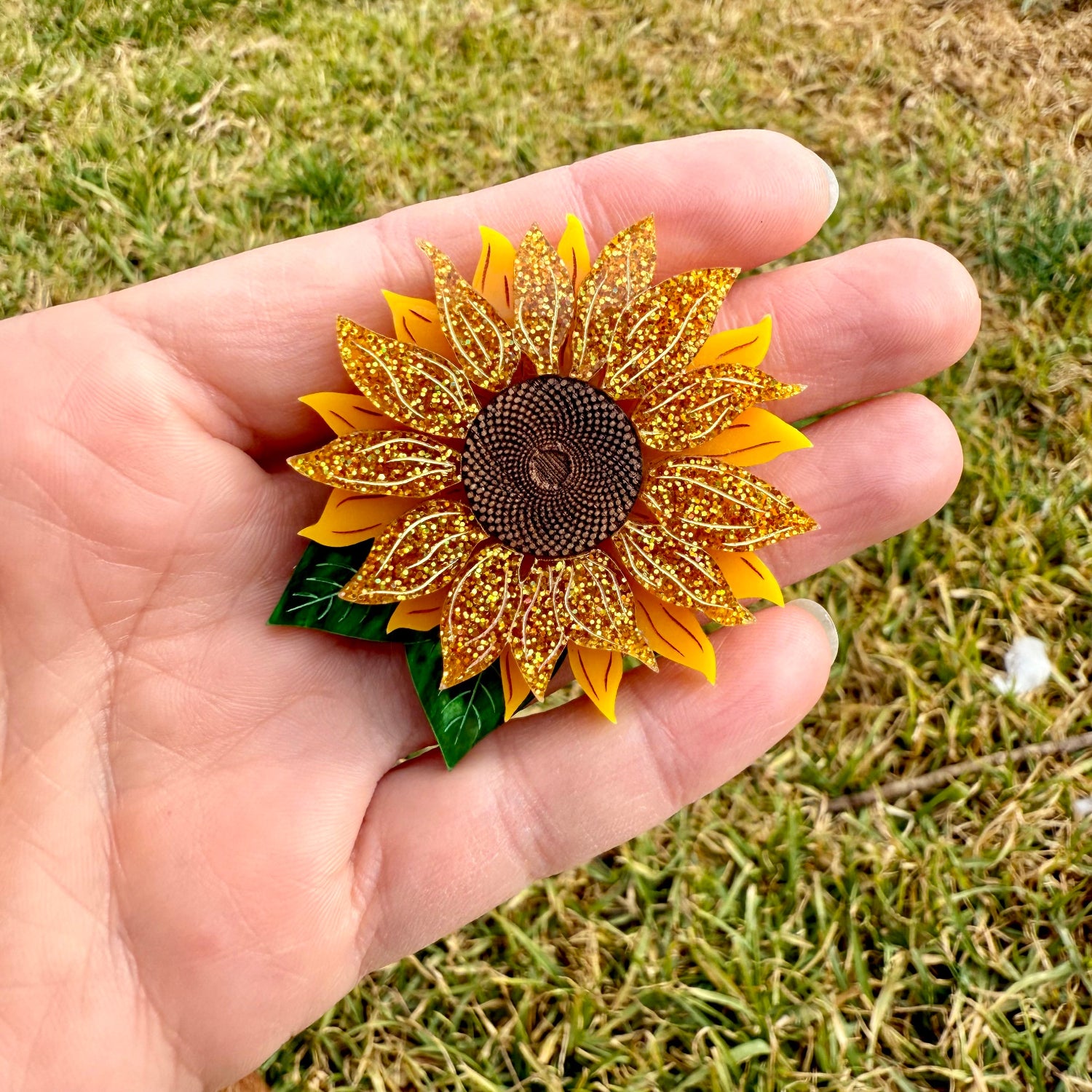 Decorative sunflower pin held in a hand with grass in the background