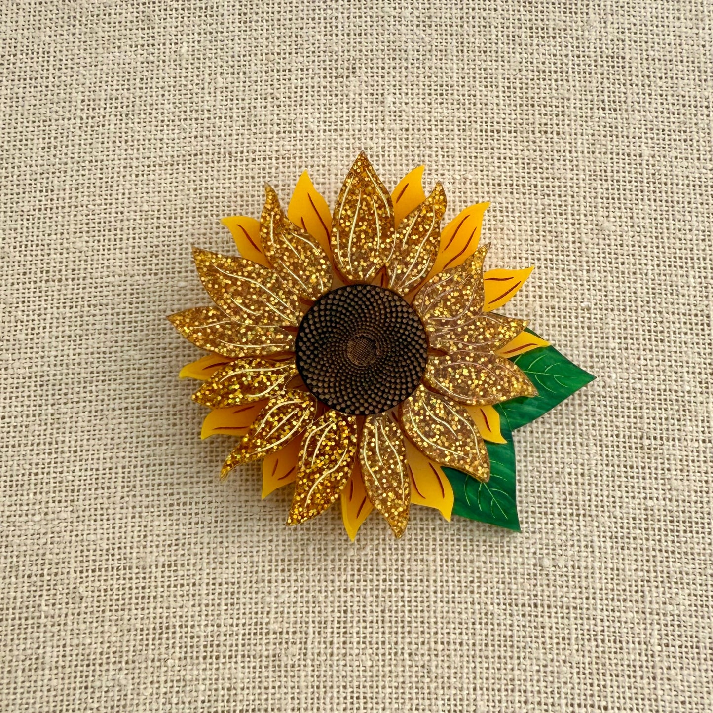 Acrylic sunflower brooch with gold petals and green leaf on a textured beige background