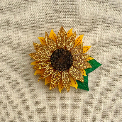 Acrylic sunflower brooch with gold petals and green leaf on a textured beige background