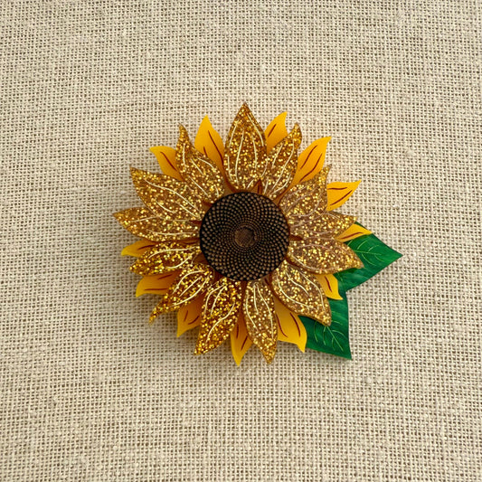 Acrylic sunflower brooch with gold petals and green leaf on a textured beige background