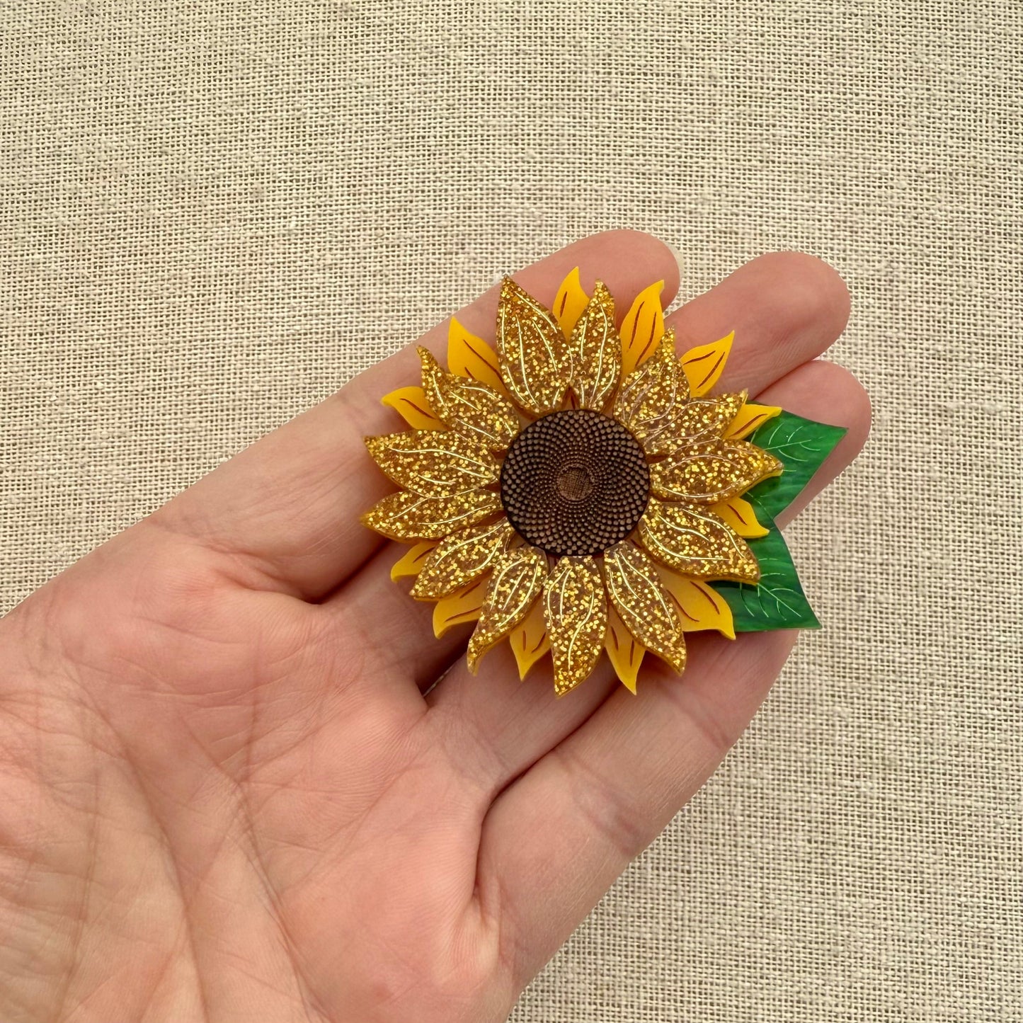 Sunflower brooch with glittery petals held in a hand against a beige background