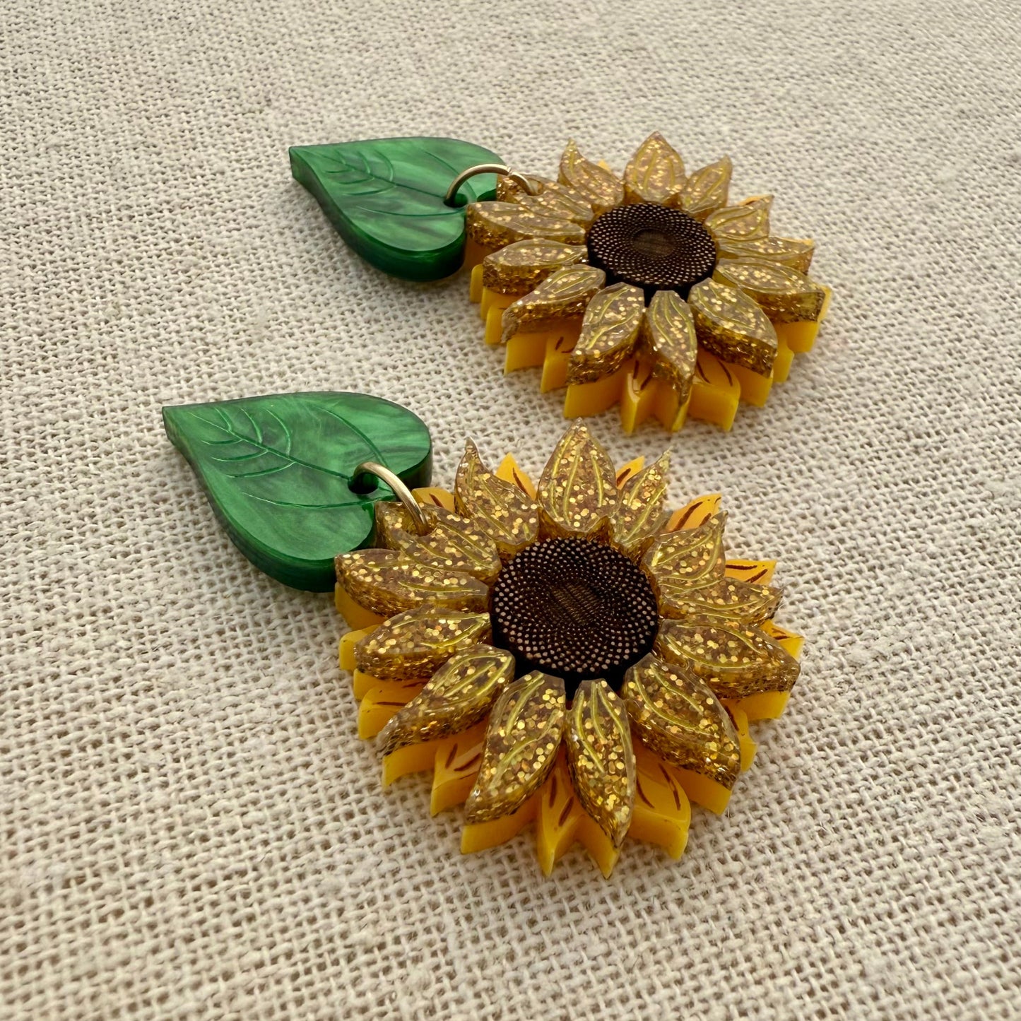 Gold sunflower-shaped earrings with green leaves on a textured beige background
