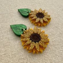 Load image into Gallery viewer, Gold sunflower-shaped earrings with green leaves on a textured beige background
