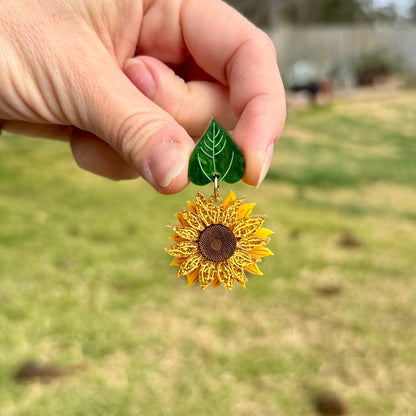 Hand holding a sunflower-shaped earring with a green leaf accent against a blurred outdoor background.