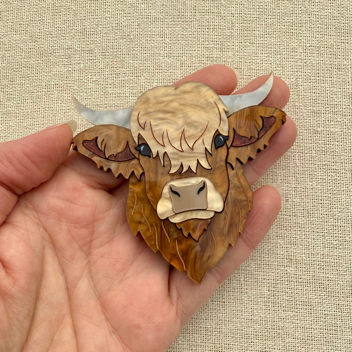 Highland cow head acrylic brooch held in a hand against a beige fabric background