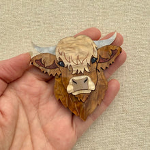 Load image into Gallery viewer, Highland cow head acrylic brooch held in a hand against a beige fabric background
