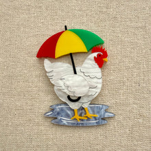 Load image into Gallery viewer, Acrylic brooch of a chicken holding a colourful umbrella, standing in a puddle on a textured beige background

