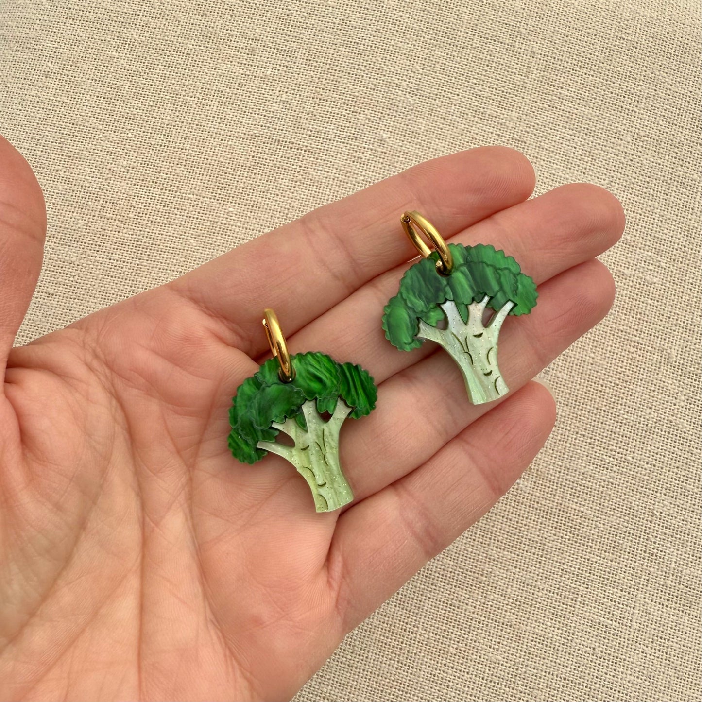 Broccoli-shaped earrings in hand against a beige fabric background.
