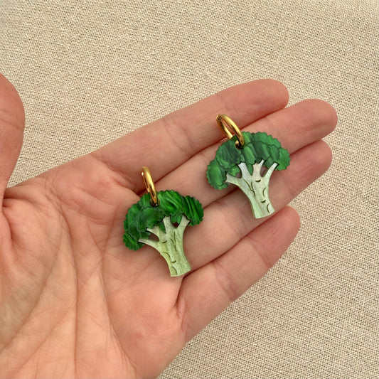 Broccoli-shaped earrings in hand against a beige fabric background.