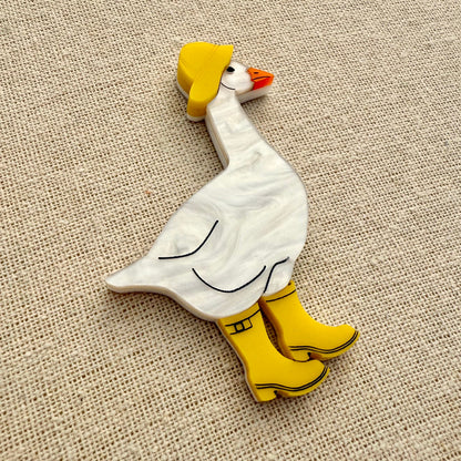 Acrylic goose brooch wearing a yellow hat and boots on a textured beige background