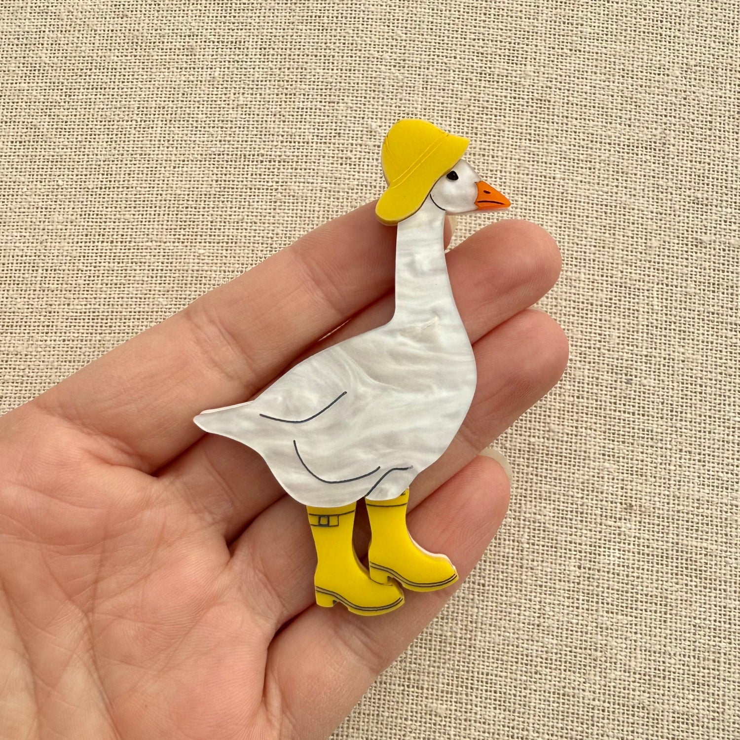 Acrylic goose brooch wearing a yellow hat and boots on a textured beige background