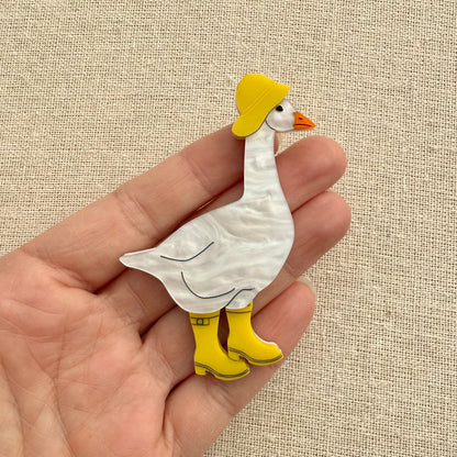 Acrylic goose brooch wearing a yellow hat and boots on a textured beige background